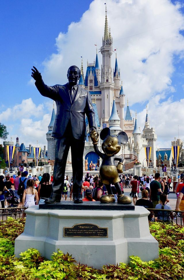 A bronze statue of Walt Disney and Mickey Mouse holding hands on a pedestal in front of a large fairytale castle with crowds of people.