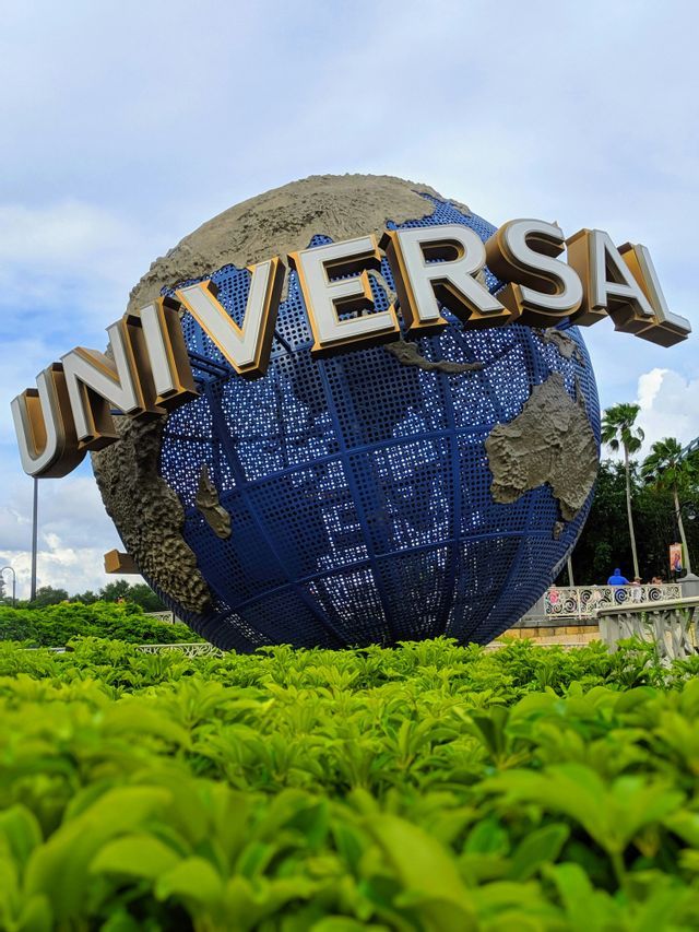 The large blue Universal Studios globe sign seen from a low angle over green hedges under a cloudy sky.