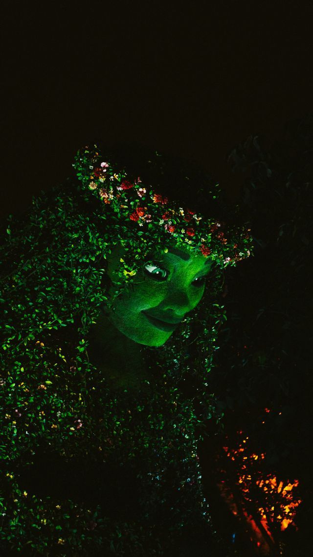 A close-up of an illuminated figure made of green leaves, wearing a crown of colorful flowers against a dark background.