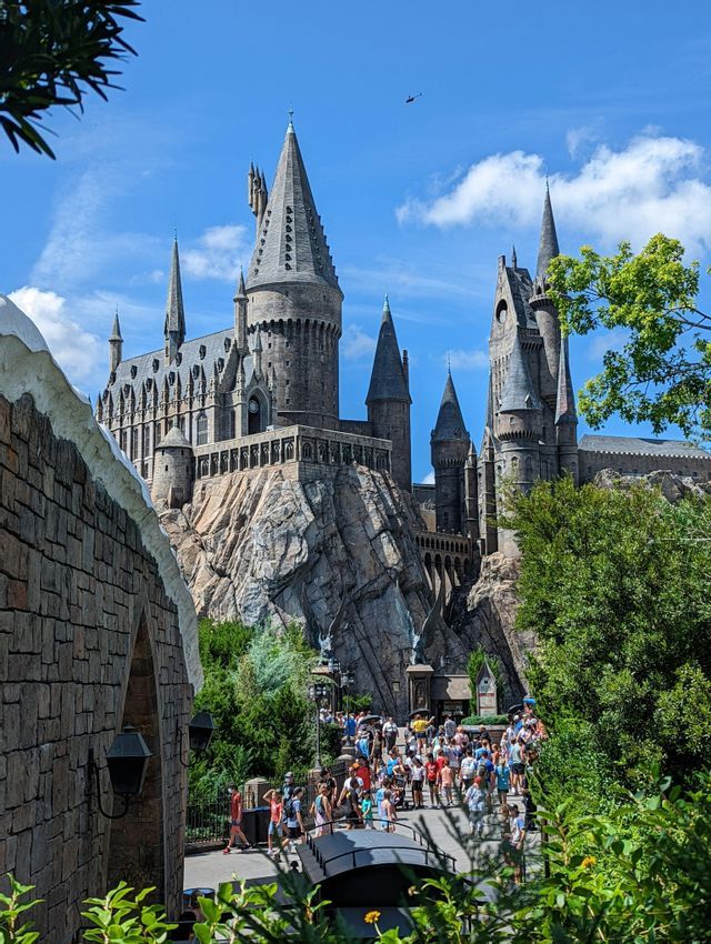 A view of Hogwarts castle perched on a rocky cliff, with a crowd of people walking on the path below under a blue sky.