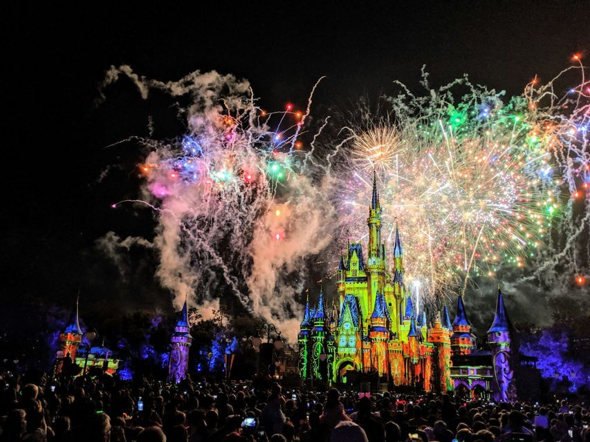A large crowd watches a colorful fireworks display exploding in the night sky over an illuminated fairytale castle.