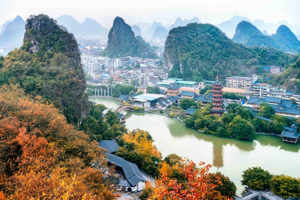 Vue aérienne d'une ville avec une pagode rouge au bord d'une rivière, entourée de montagnes karstiques, avec des arbres d'automne colorés au premier plan.