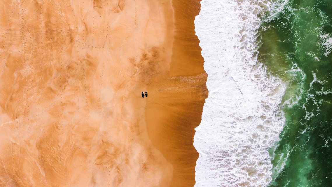 Vue aérienne de deux personnes sur une vaste plage de sable orange, près de l'océan vert mousseux.