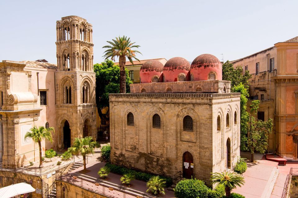 Una storica chiesa in pietra con tre cupole rosse e un alto campanile in un cortile con palme.