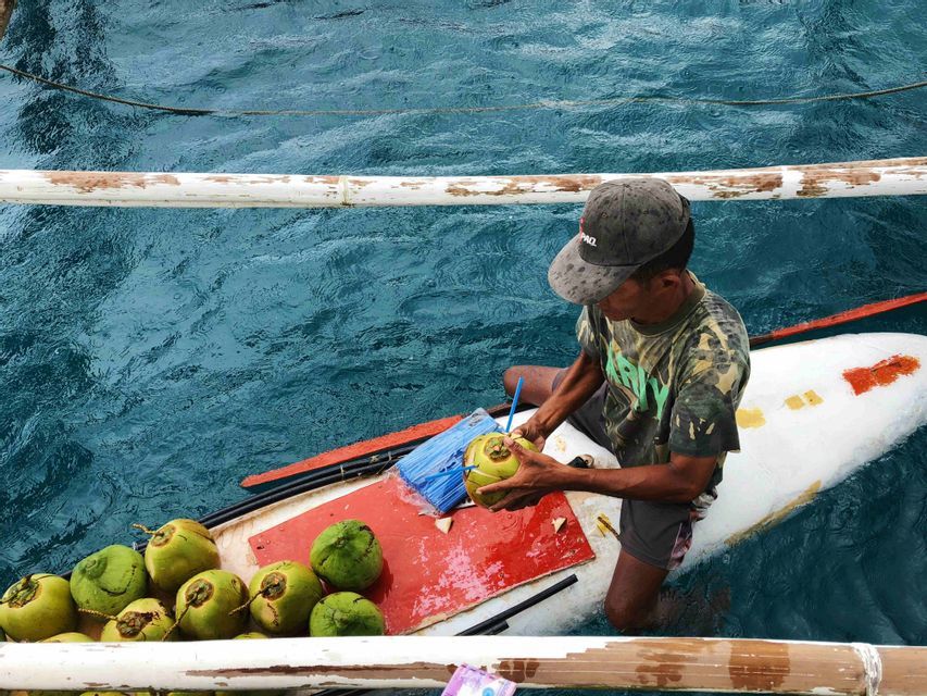 Un uomo su una piccola barca prepara una noce di cocco fresca e verde con una cannuccia, circondato da altre noci di cocco sull'acqua blu.