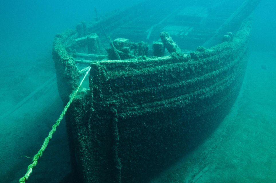 Un grande relitto di legno, coperto di alghe, giace sul fondale marino con una spessa corda attaccata alla sua prua.