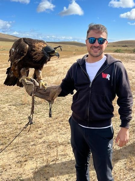 Un uomo con occhiali da sole sorride mentre tiene una grande aquila incappucciata su un guanto di cuoio in un paesaggio arido e collinare.