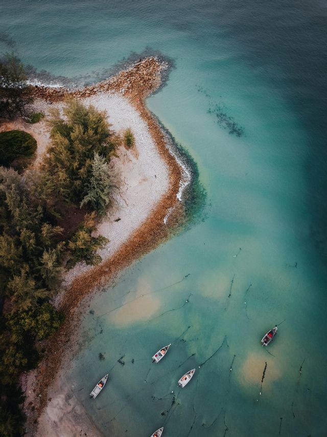 Luftaufnahme von oben: Eine geschwungene Küstenlinie mit einem Kiesstrand, Bäumen und mehreren kleinen Booten, die auf klarem, türkisfarbenem Wasser schwimmen.