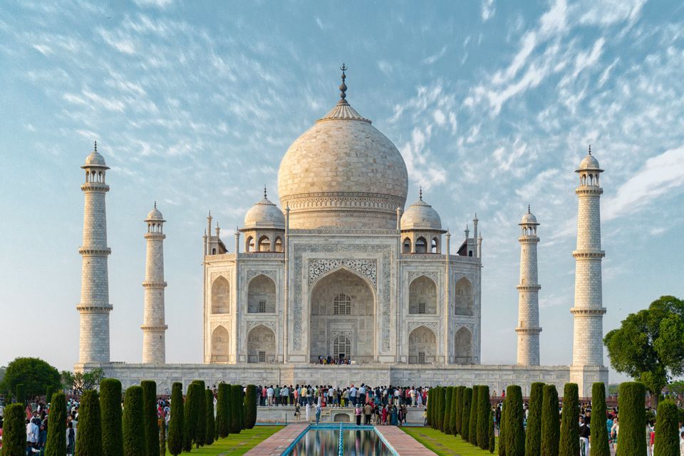 Una vista frontale del Taj Mahal con turisti riuniti davanti, affiancato da cipressi e uno specchio d'acqua in primo piano.