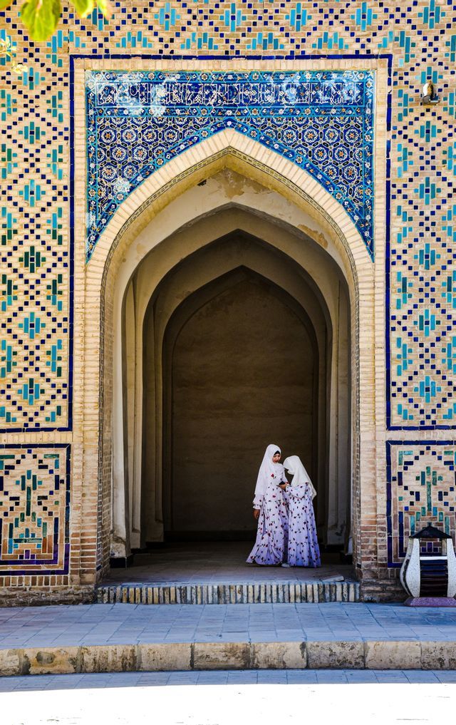 Due giovani ragazze in hijab bianchi e abiti coordinati si trovano in un arco in mattoni ornato decorato con piastrelle a mosaico blu.