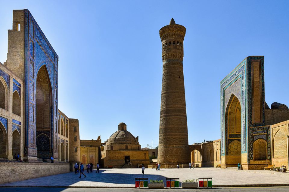 Un alto minareto in mattoni ed edifici decorati con mosaici blu che circondano una piazza soleggiata dove le persone passeggiano.