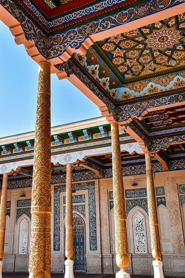 Colonne in legno riccamente intagliate sostengono un portico con un soffitto colorato e decorato, sotto un cielo azzurro sereno.