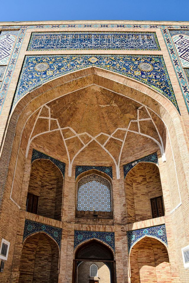 Vista dal basso di un sontuoso edificio in mattoni con un grande arco a volta, decorato con mosaici di piastrelle blu e gialle sotto un cielo sereno.