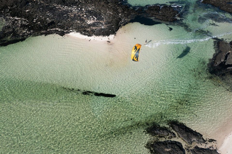 Vue aérienne d'une personne faisant du kitesurf dans un lagon clair avec des plages de sable blanc et des roches volcaniques sombres.