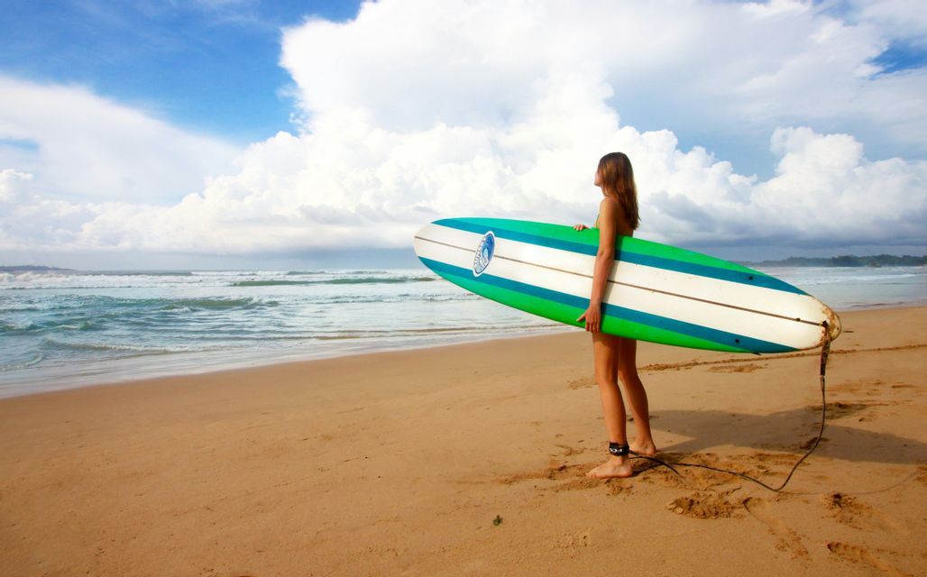 Une femme tient une planche de surf sur une plage de sable, regardant l'océan.