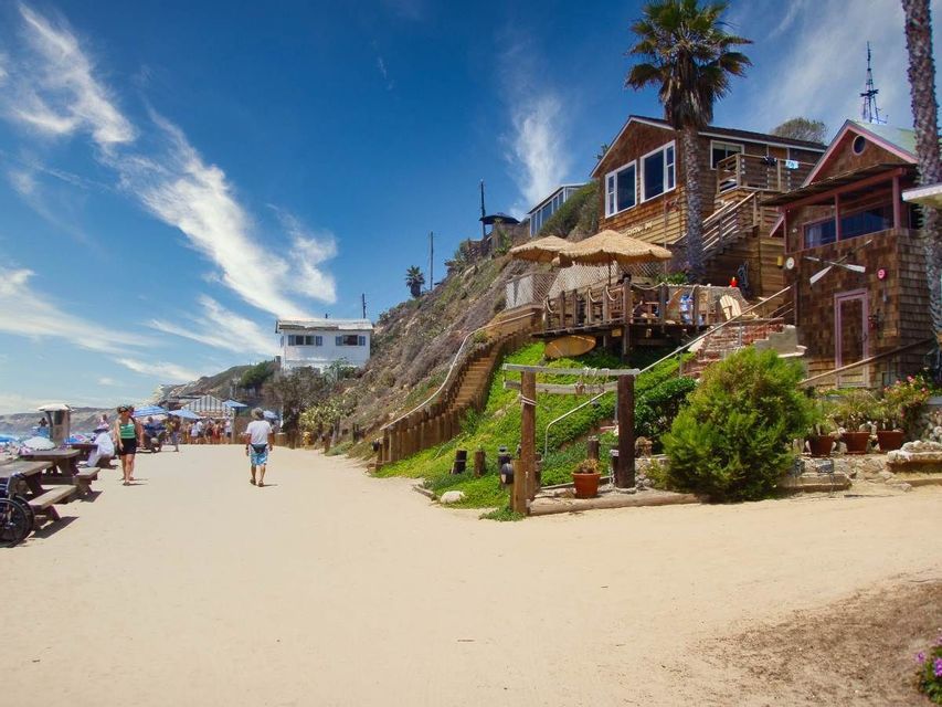 Le persone camminano lungo un sentiero sabbioso accanto a case sulla spiaggia in legno costruite su una collina sotto un cielo soleggiato e azzurro.