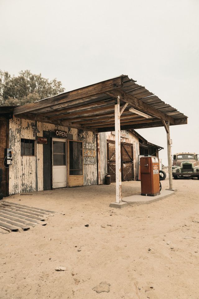 Una stazione di servizio rustica e invecchiata con una pompa di benzina arancione d'epoca e un vecchio camion parcheggiato su terreno sabbioso.