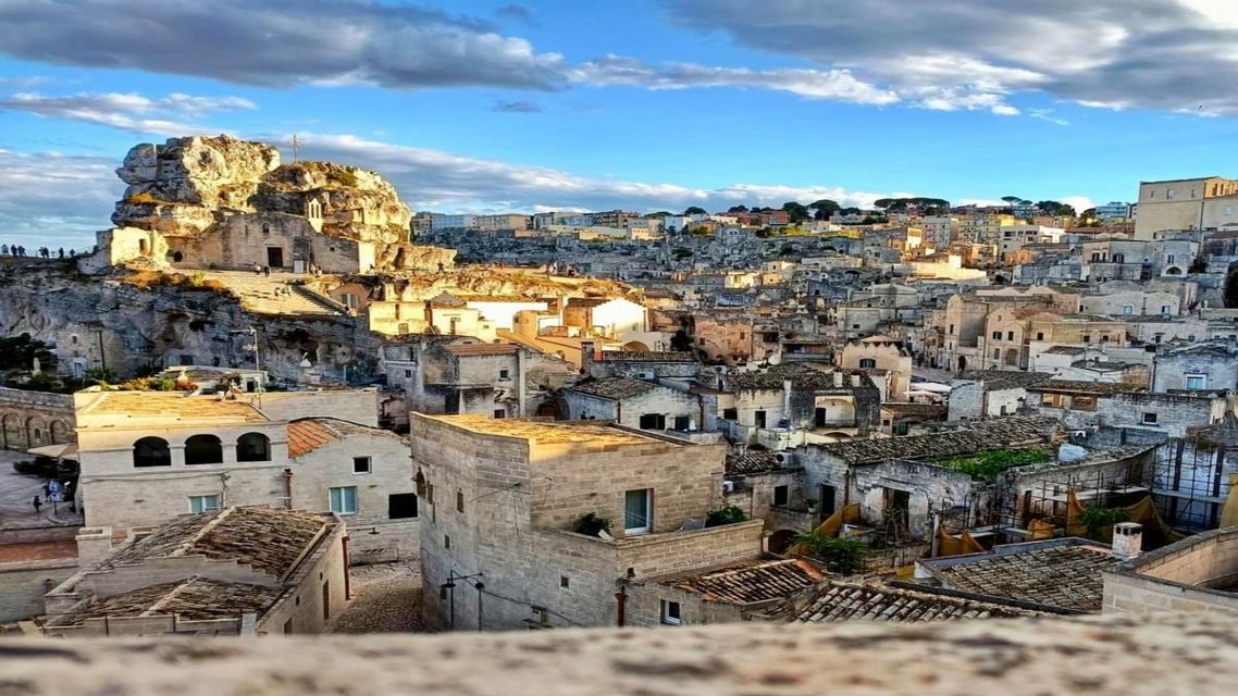 Una grande formazione rocciosa con una chiesa domina una storica città di edifici in pietra all'ora d'oro sotto un cielo parzialmente nuvoloso.