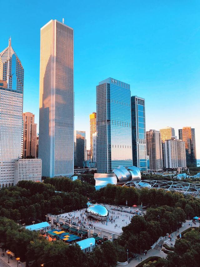 An elevated view of a city park with a large, reflective bean-shaped sculpture, surrounded by skyscrapers under a clear blue sky.