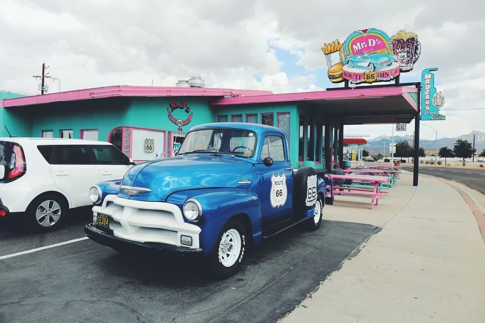 A vintage blue pickup truck with a Route 66 logo is parked in front of a colorful, retro American diner on a cloudy day.
