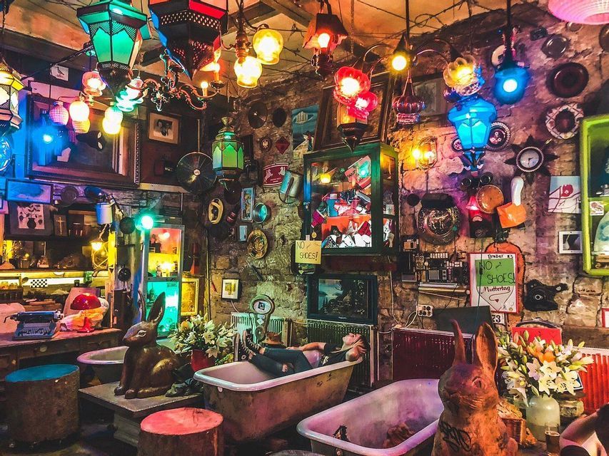 A person relaxes in a bathtub inside an eclectic room filled with colorful hanging lanterns and quirky objects.