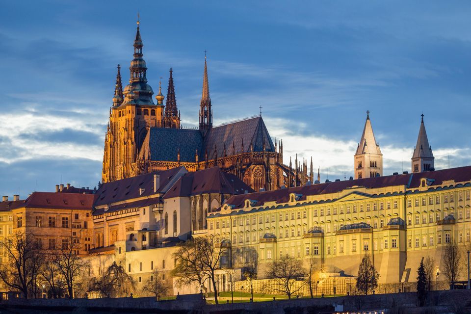 A large, illuminated Gothic cathedral with spires stands over a historic building complex at twilight.
