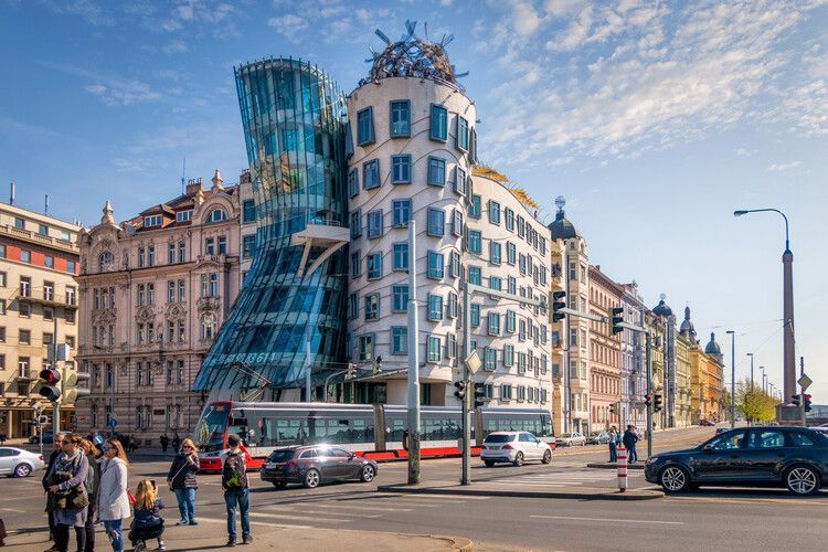 The modern Dancing House building, with its curved glass facade, stands on a busy city corner with cars, pedestrians, and a tram.