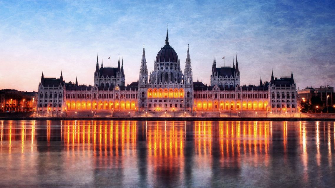 An ornate, illuminated building with a central dome on a riverbank at dusk, its lights reflecting in the still water.
