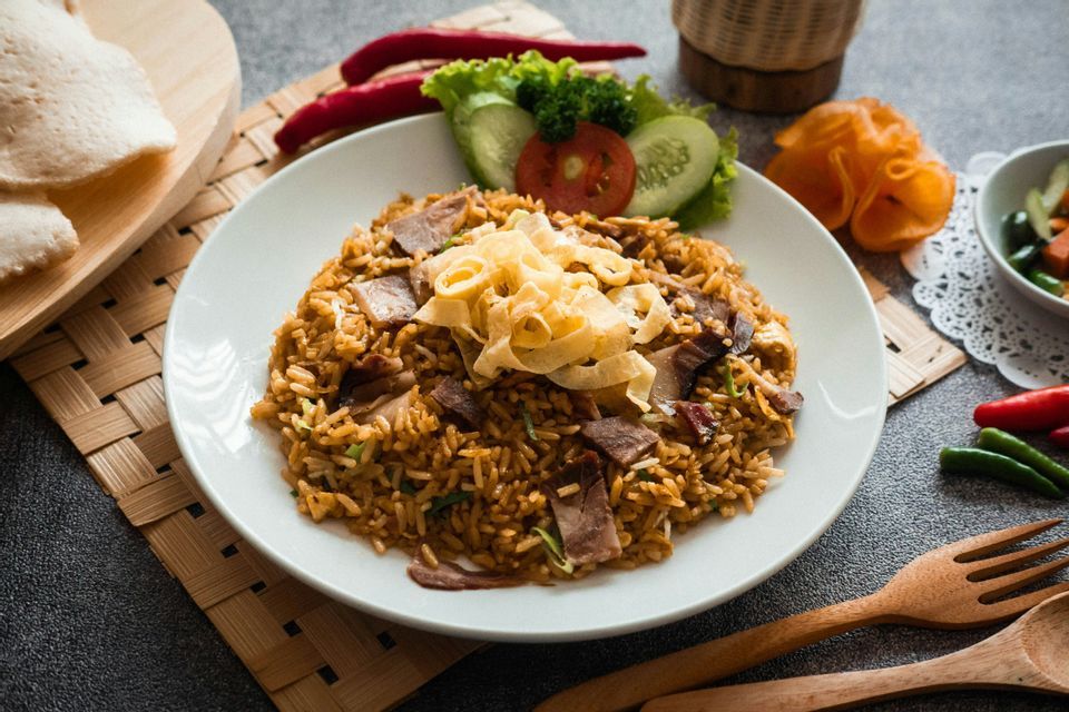 Un piatto di Nasi Goreng con carne e frittata sminuzzata, servito con verdure fresche, cracker e peperoncini su un tavolo scuro.