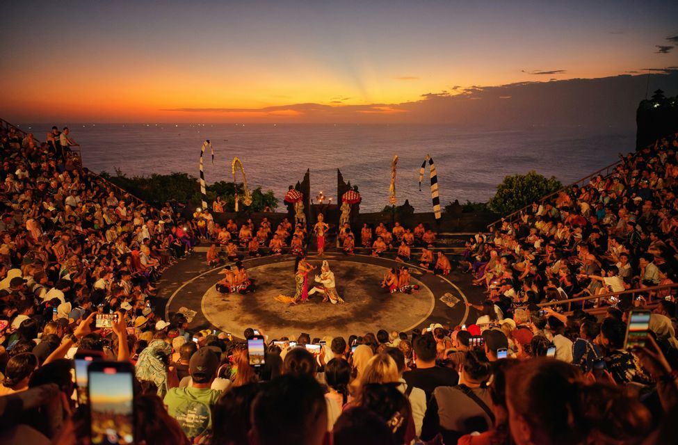 Una grande folla assiste a uno spettacolo su un palco circolare in un anfiteatro sulla scogliera affacciato sull'oceano al tramonto.