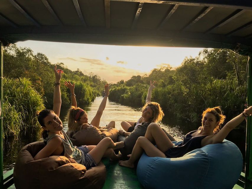 Un viaje en grupo de WeRoad de mujeres relajándose en pufs en un barco navegando por un río selvático al atardecer.