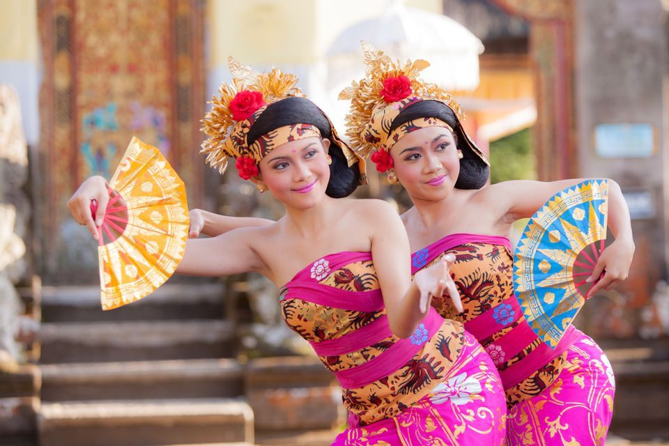 Dos mujeres con trajes tradicionales ornamentados y tocados dorados realizan un baile sincronizado sosteniendo abanicos coloridos.