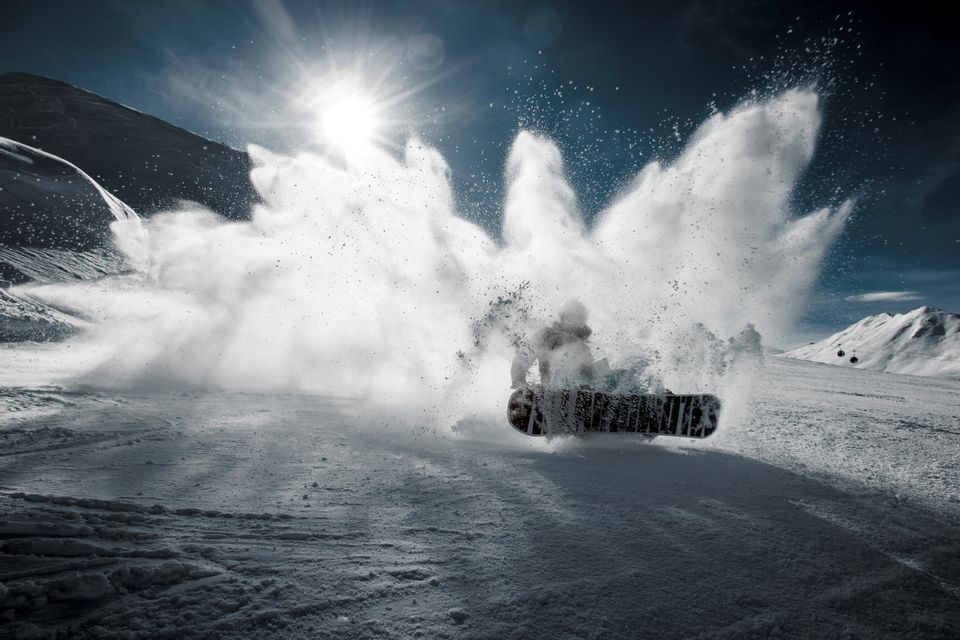 Uno snowboarder scende una pista, sollevando una grande nuvola di neve fresca sotto un sole splendente.