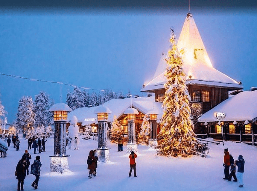 Una piazza di paese innevata al tramonto, con edifici illuminati, un grande albero di Natale acceso e persone che camminano nella neve.