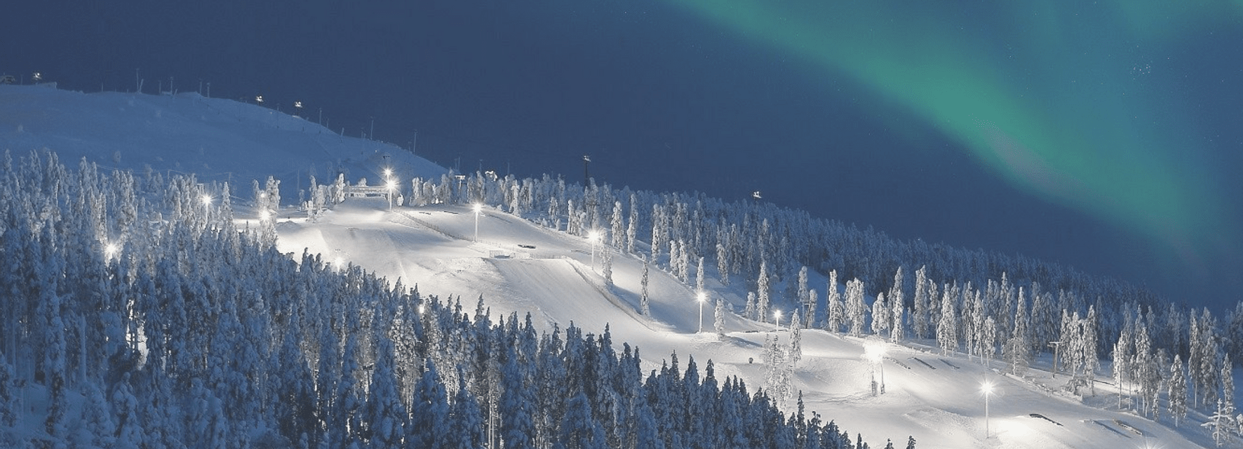 Una pista da sci illuminata si snoda attraverso una foresta innevata di notte, con l'aurora boreale che brilla nel cielo blu scuro.