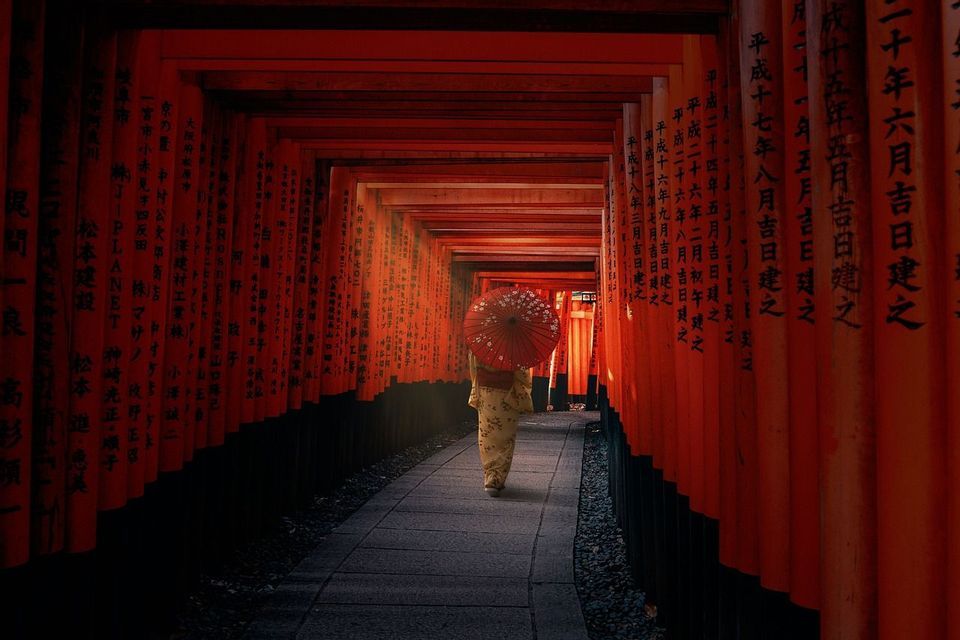 Una persona in kimono tiene un ombrello rosso mentre cammina lungo un sentiero di pietra attraverso un tunnel di torii rossi.