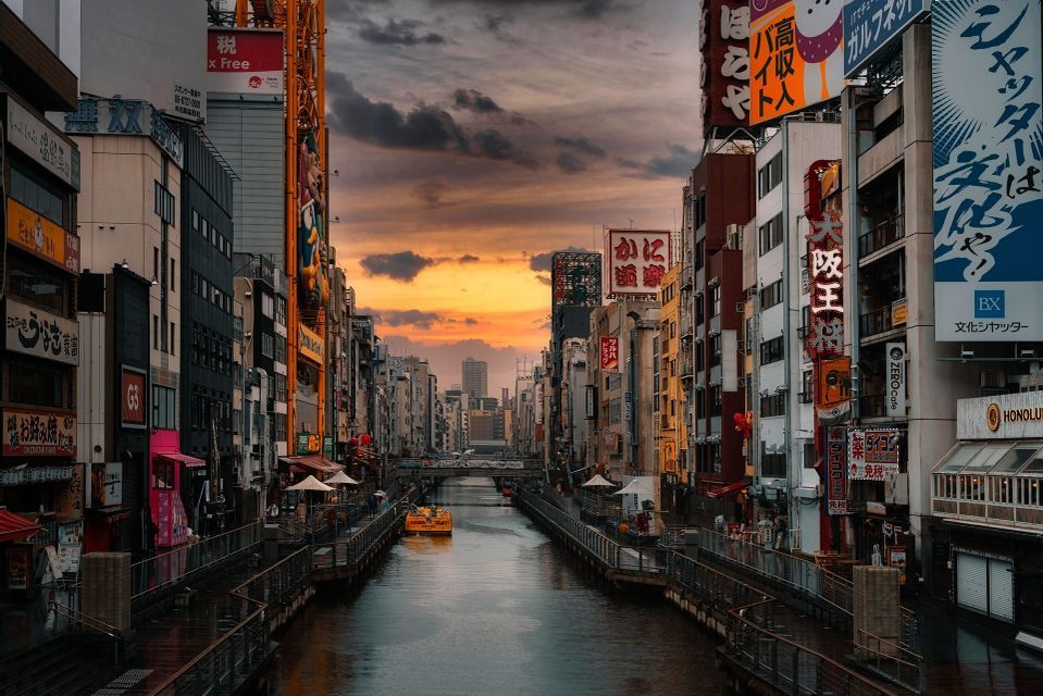 Una barca gialla naviga lungo un canale circondato da edifici con insegne e cartelloni giapponesi, il tutto sotto un cielo al tramonto colorato.