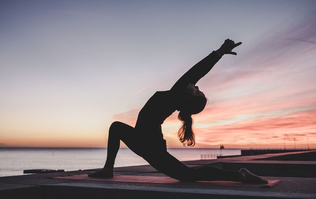 La silhouette di una persona in posizione yoga dell'affondo su un tappetino, con lo sguardo rivolto al mare durante un tramonto colorato.