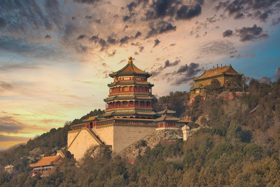 Una pagoda cinese a più livelli si erge su una collina boscosa con altri templi sotto un drammatico cielo al tramonto.