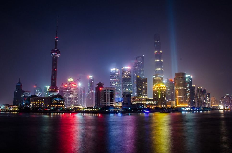 Un ampio skyline cittadino di notte, con edifici illuminati e una torre che riflette luci colorate sull'acqua.
