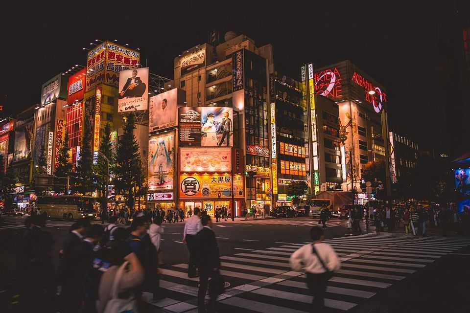 Persone che attraversano una strada cittadina trafficata di notte, con alti edifici illuminati da insegne al neon colorate e grandi cartelloni pubblicitari.