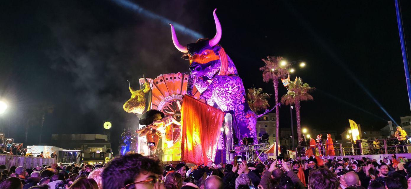 Un grande e sontuoso carro allegorico a forma di toro, illuminato da luci viola, si muove attraverso una folla fitta durante un festival notturno.