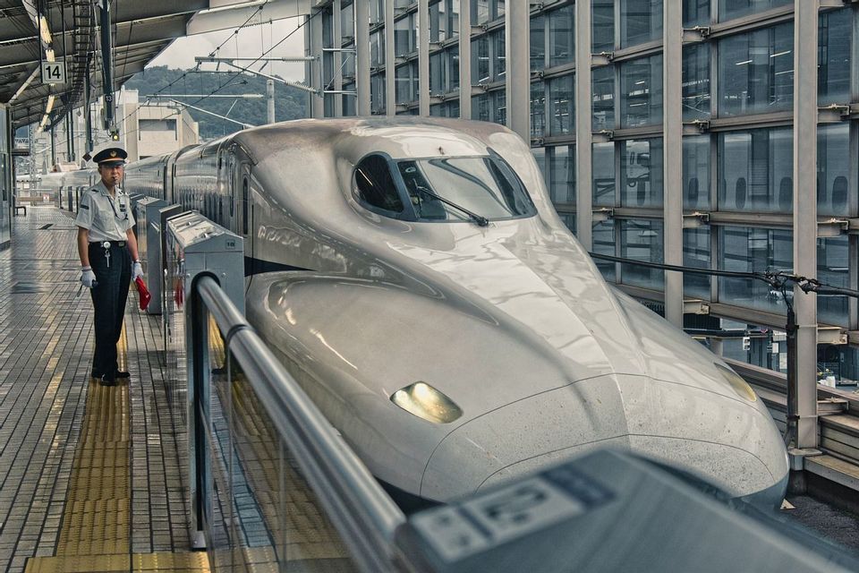 Un capotreno in uniforme si trova accanto a un treno proiettile Shinkansen su una moderna piattaforma ferroviaria.