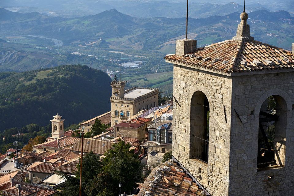 Una vista dall'alto di un borgo storico con campanile in pietra e tetti in terracotta, affacciato su una valle con dolci colline verdi.