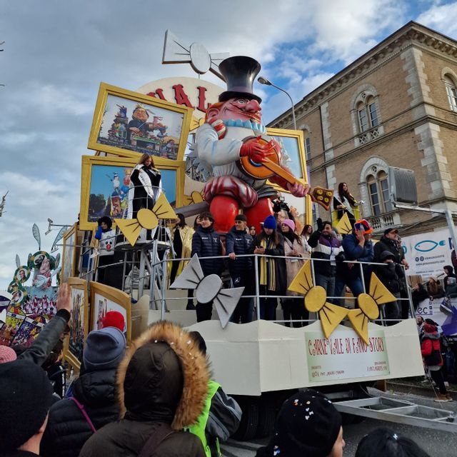 Un grande carro di carnevale con una gigantesca figura caricaturale che suona la chitarra è osservato da una folla di persone durante una sfilata di strada.