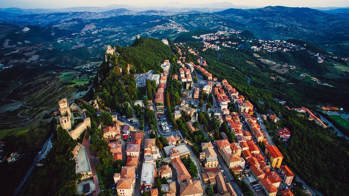 Vista aerea di un borgo storico con tetti in terracotta e una fortezza su un crinale montuoso e boscoso.