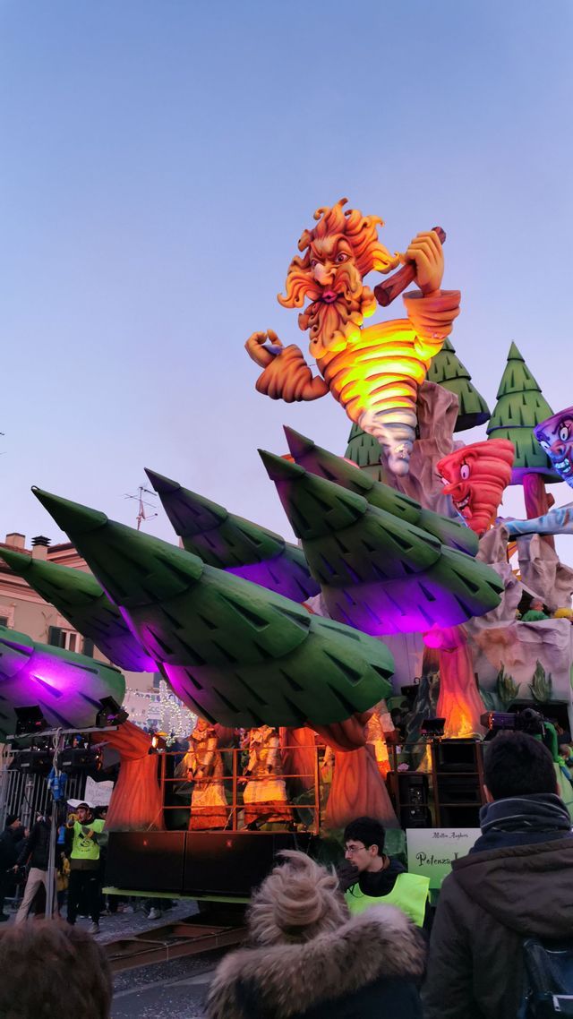Un grande e colorato carro di carnevale con una gigantesca figura di tornado a cartone animato e alberi stilizzati osservato da una folla al crepuscolo.