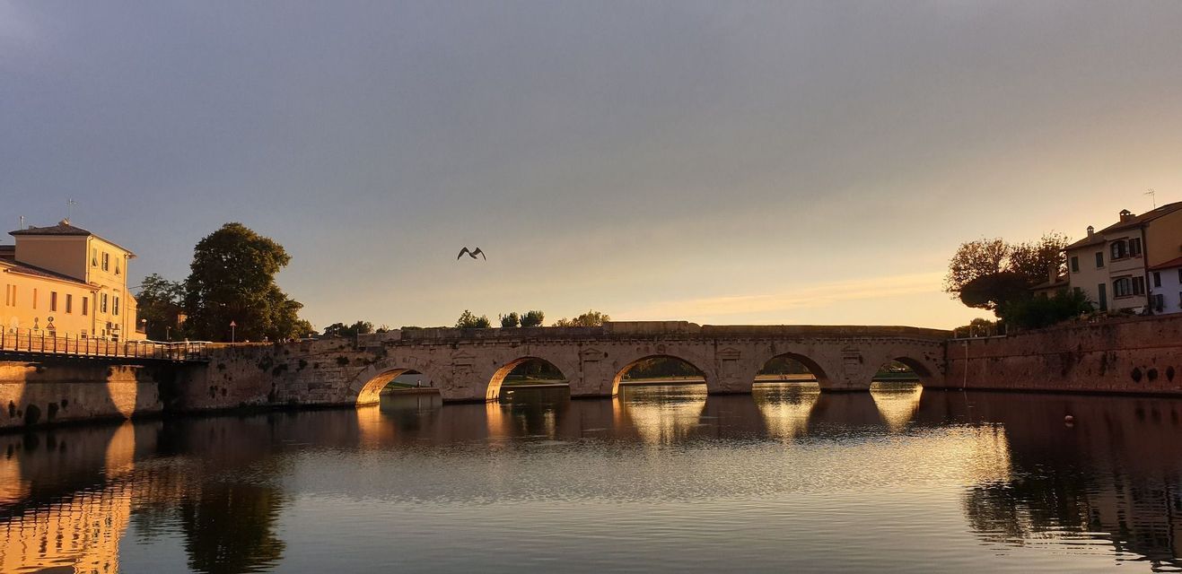 Un ponte in pietra a più arcate che attraversa un fiume calmo al tramonto, con il suo riflesso visibile nell'acqua e un uccello che vola sopra.