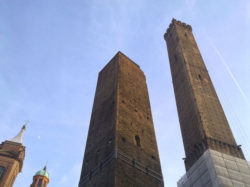 Una veduta dal basso di due alte e storiche torri in mattoni che si ergono contro un cielo azzurro chiaro, con impalcature visibili alla base di una.