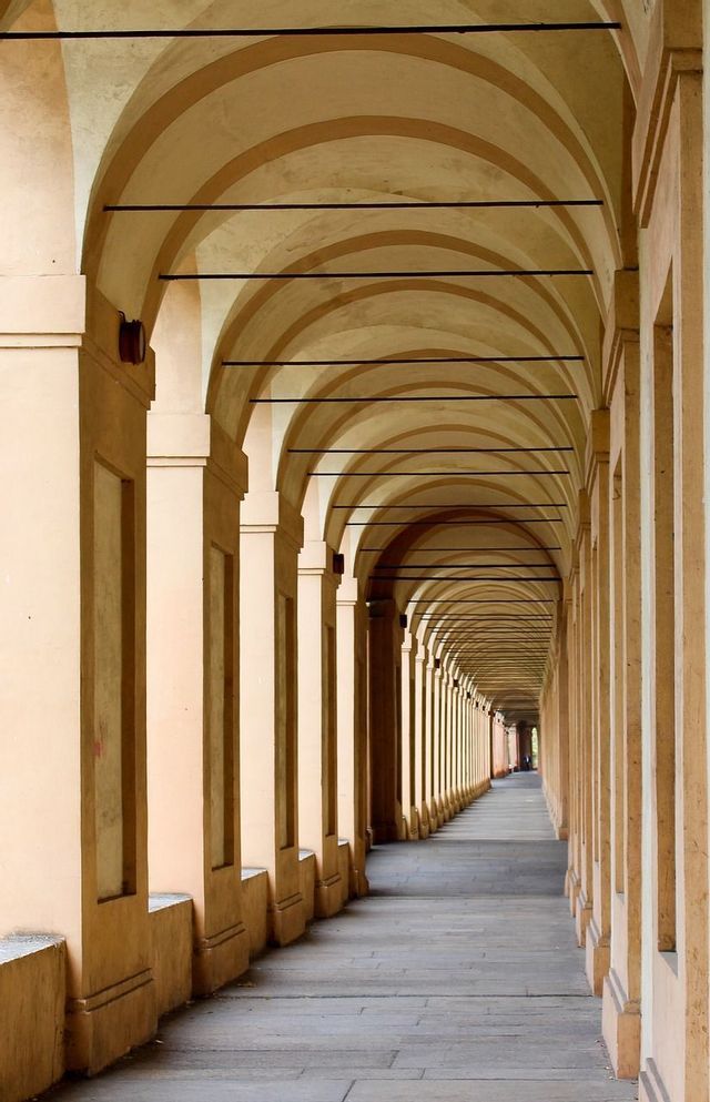 Un lungo portico con pilastri beige ripetuti e archi a volta a strisce, che creano un punto di fuga lungo una passerella in pietra.